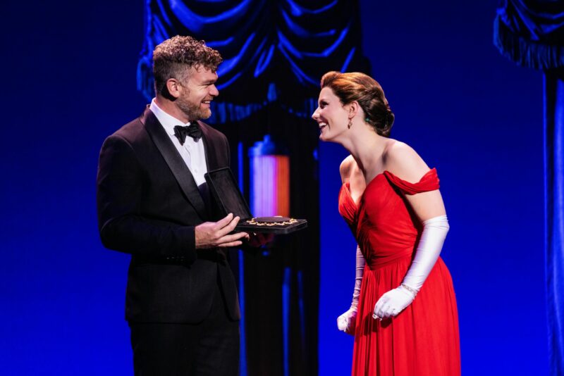 Chase Wolfe and Ellie Baker in Pretty Woman the Musical. Edward, dressed in a tux, holds a necklace in a jewelry box out to Vivian who happily looks at Edward.