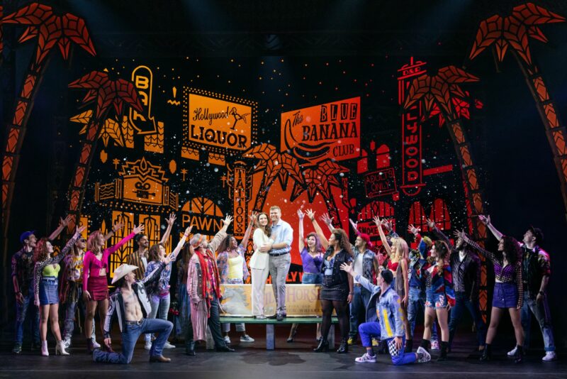 The cast of Pretty Woman the Musical. Vivian and Edward stand center on a bench holding each other. A group of people surround them holding one arm out to them. There are multiple neon signs behind them.