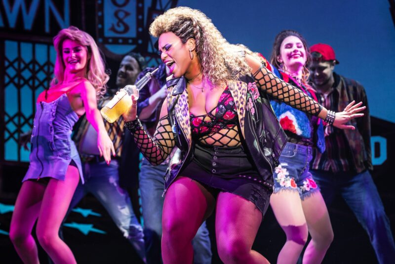 Rae Davenport and The Company of Pretty Woman: The Musical. A woman, wearing a short black skirt, fishnet top, and leather vest, sings into a bottle of alcohol. A group of people dance behind her.