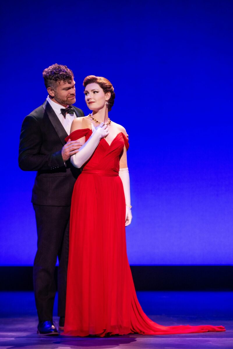 Chase Wolfe and Ellie Baker in Pretty Woman the Musical. Edward, dressed in a tux, stands behind Vivian who wears a long elegant red dress and white gloves. Vivian puts her hand up to the necklace she wears.